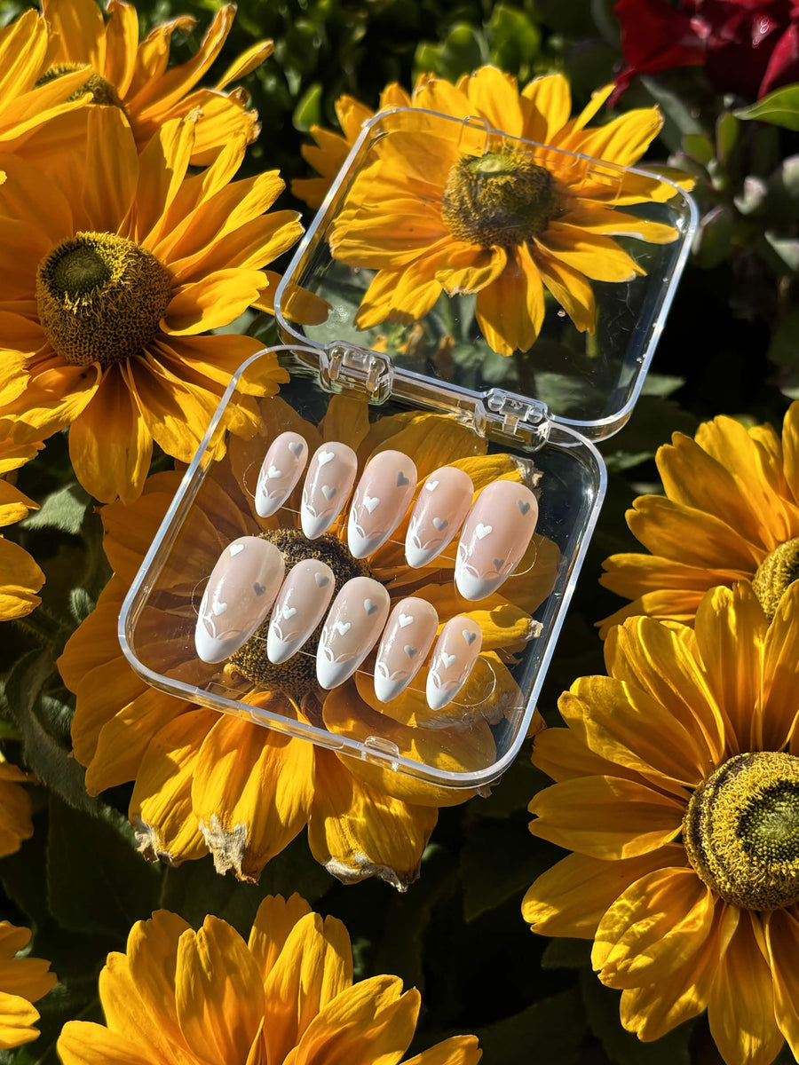 Blushing Hearts press-on nails — ten almond nails with white French tips, hand-painted hearts and fine white line-work on a milky nude base, in an open clear case